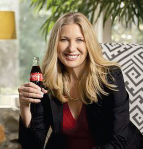 Dagmar Boggs Smiling in a black suit and red top holding a glass bottle of Coca-Cola, seated indoors with natural light and stylish modern decor in the background.