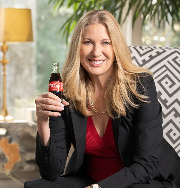 Dagmar Boggs Smiling in a black suit and red top holding a glass bottle of Coca-Cola, seated indoors with natural light and stylish modern decor in the background.