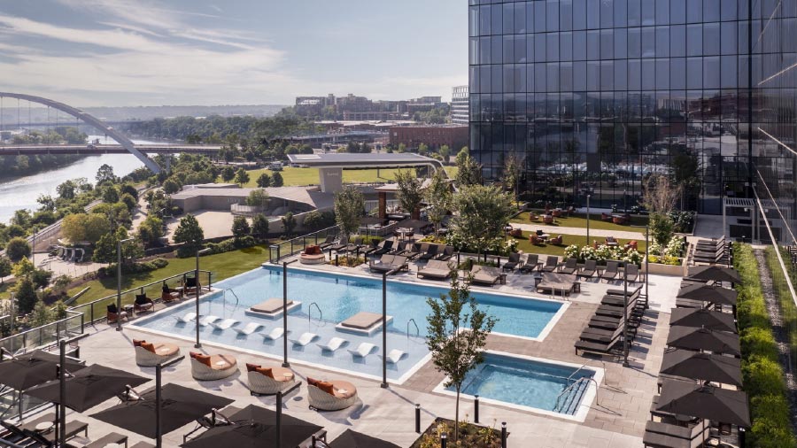 Scenic view of the 7th-floor terrace at the Four Seasons Hotel Nashville, featuring a modern outdoor pool area with sun loungers, circular cabanas, and sleek black umbrellas. The terrace overlooks the Cumberland River and city skyline, with a nearby pedestrian bridge and lush green spaces enhancing the upscale urban atmosphere.